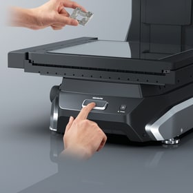 A finger presses the start button on the device's base after placing the part.